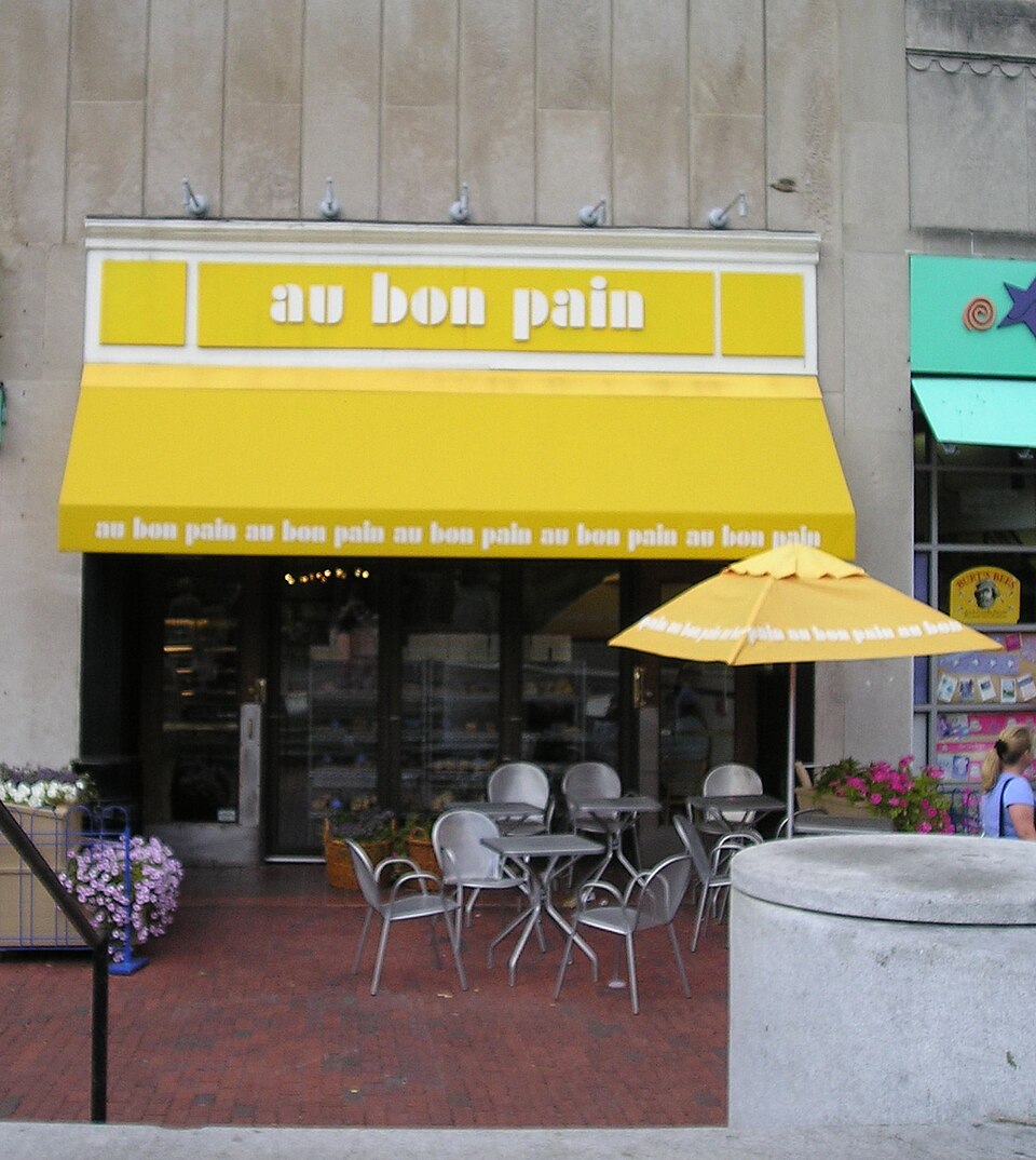 The Au Bon Pain storefront on Brattle Street, a yellow awning and sign over the entrance, outdoor metal tables and chairs on the brick pavement in front.