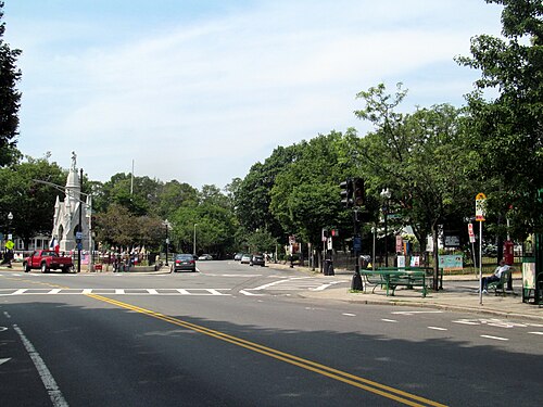 The intersection at Centre and South Streets, the Soldier's Monument behind a picket fence, a green bus stop bench and traffic signals over the crosswalk.