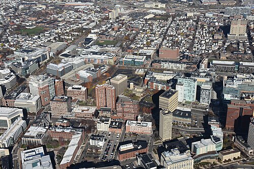 An aerial view over Kendall Square showing brick mid rise buildings, glass towers, parking lots and the grid of streets running toward the Charles River.
