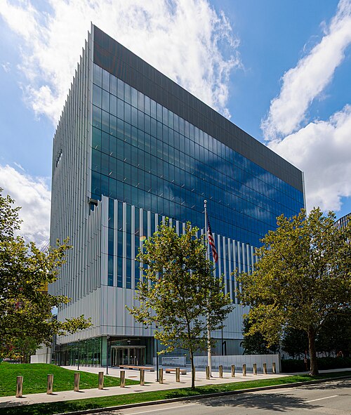 A tall glass office tower with vertical white louvers, a flag out front and a low glass entrance pavilion at street level.