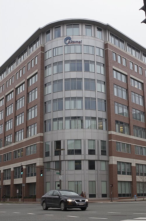 A brick office building with a rounded glass and metal corner bay carrying the Akamai sign, traffic lights and a car at the intersection below.