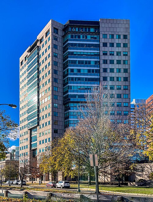 A stone faced office tower with narrow windows in two tones of granite, parked cars and trees at its base under a clear sky.