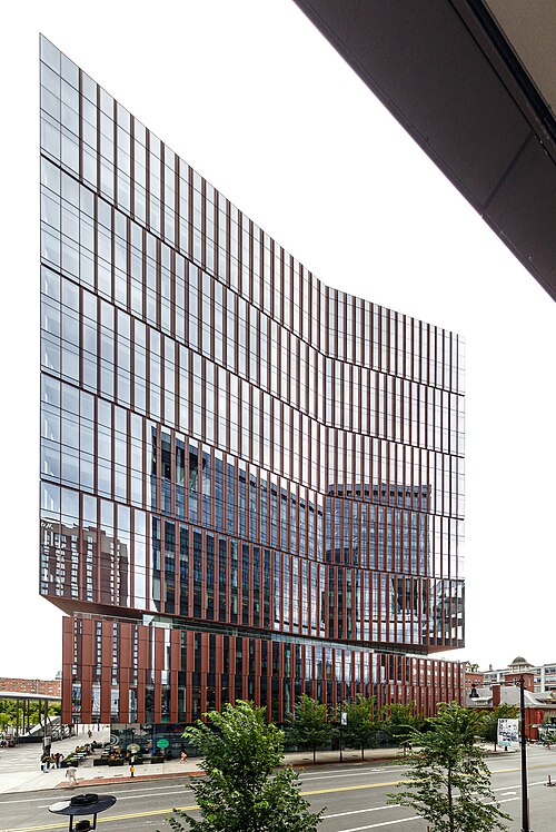 A glass tower with an angled, faceted facade in copper toned vertical fins, rising above street trees.