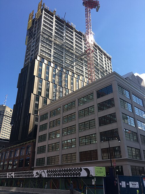 A glass and metal panel tower under construction with a tower crane, rising directly behind a lower brick mill building with tall multi pane windows.
