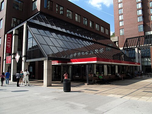 The glass and steel headhouse of the Kendall/MIT T station, benches along the front under a red awning band, the MIT Coop sign on the building behind.