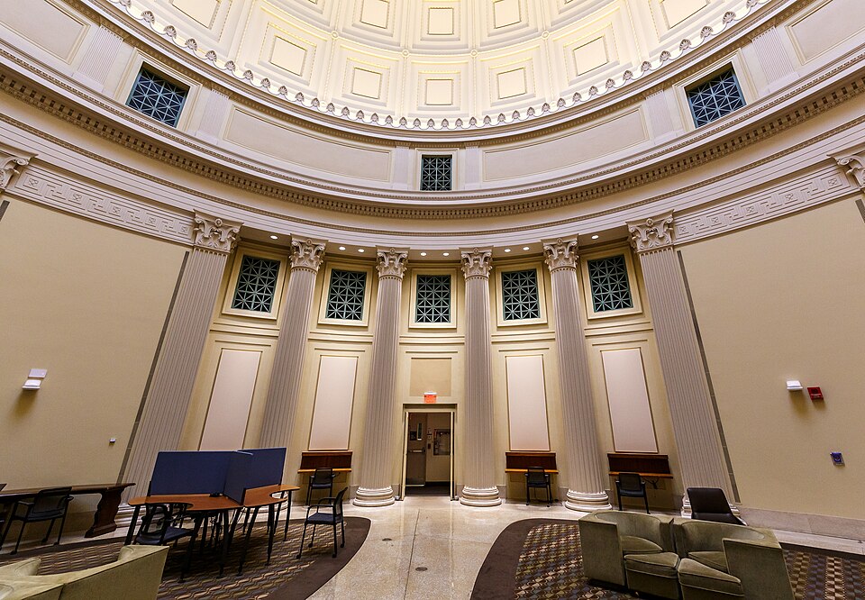 The semicircular colonnaded reading room under the Great Dome, Corinthian columns and arched clerestory windows, study desks and a clock on the far wall.