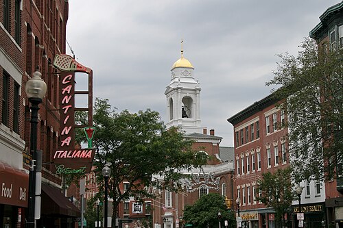 Hanover Street on a festival day, a neon Cantina Italiana sign on a bracket over the sidewalk, brick buildings both sides, the gold dome and white steeple of St. Stephen's Church rising above the street.