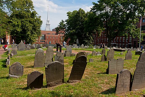 Rows of slate colonial headstones on a grass terrace, brick rowhouses and the white steeple of Old North Church visible beyond the trees.