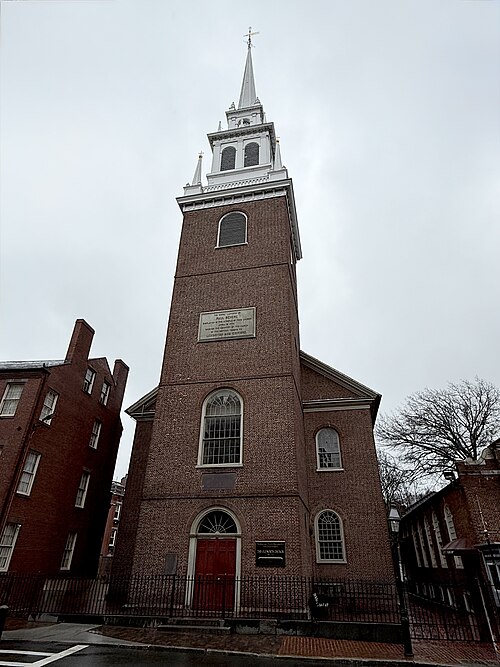 The brick front facade and white steeple of Old North Church from street level, a bronze plaque about the signal lanterns mounted on the tower, iron fence in front.