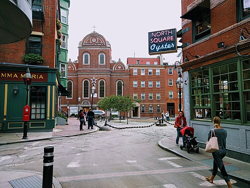 North Square, a neon North Square Oyster sign hanging over the cobbled junction, a red brick church with a gold domed cupola behind a row of brick apartment buildings, people crossing with a stroller.