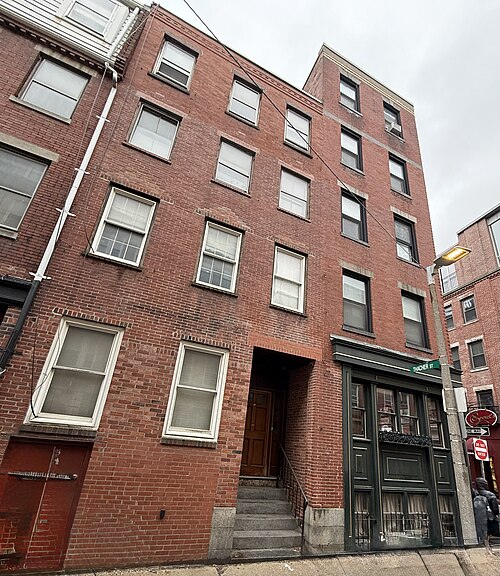A four and five storey red brick apartment building at the corner of Prince and Thacher Streets, a THACHER ST street sign mounted on the corner, a shop with dark green storefront windows at ground level.