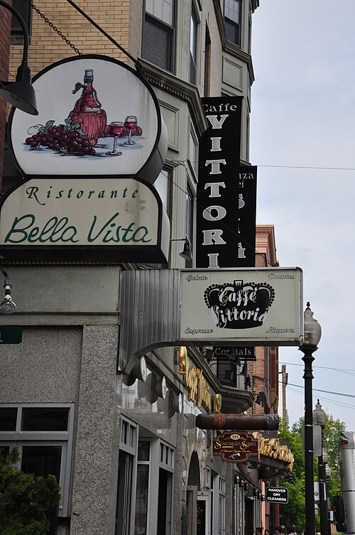 A row of restaurant and cafe signs mounted perpendicular to a brick building on Hanover Street, Ristorante Bella Vista and Caffe Vittoria signs stacked one below the next above the sidewalk, more signs continuing down the block.