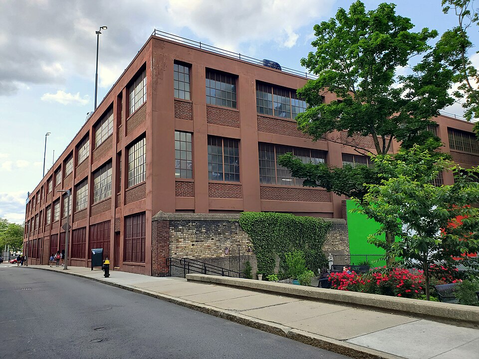 A three storey red brick parking garage on Prince Street, large multi pane windows along its side, a low stone wall and ivy at street level, a green panel leaning against the fence.