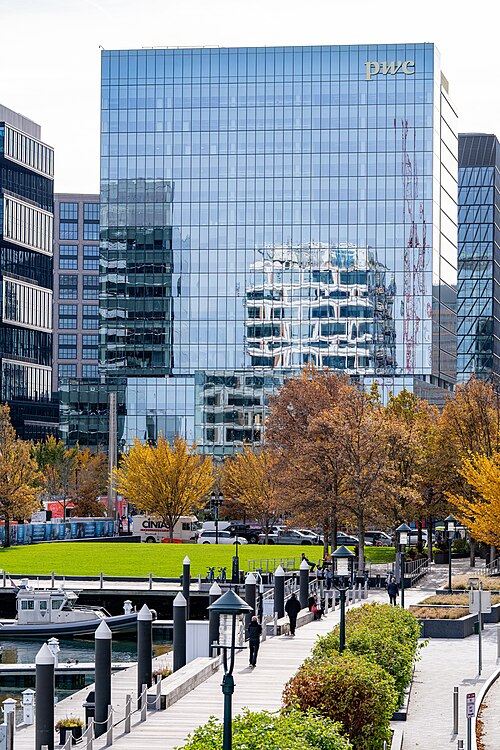 A glass office tower marked PWC rising behind a harborwalk park, autumn trees, bike racks and a marina with a small boat, people walking the boardwalk.