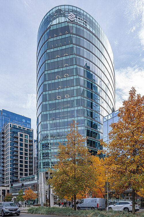 A curved glass office tower marked PTC with a peaked glass roof, seen from street level past a row of yellow autumn trees.