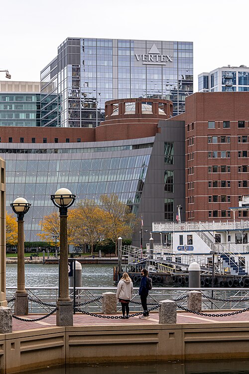 A glass tower marked VERTEX behind a domed brick rotunda and a curved glass wall, a tour boat docked in front along a railed promenade.