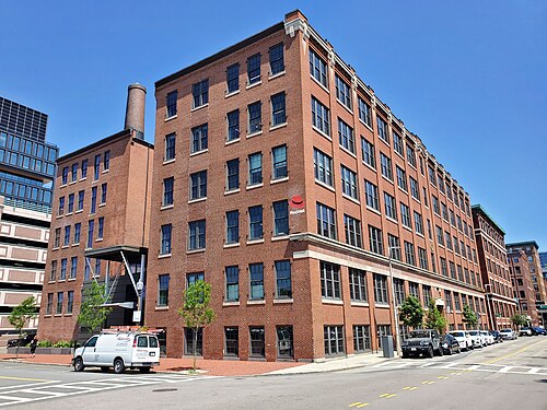 An older red brick warehouse on a street corner, five storeys with a rooftop chimney and stone trimmed window heads, a glass tower reflected in a neighboring building's windows.