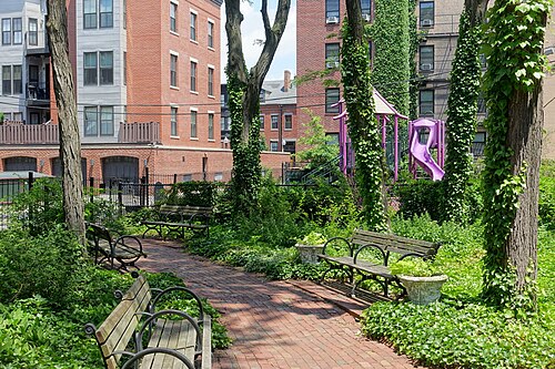 A brick path through Newland Street Park, wooden benches and ivy-covered trees, brick apartment buildings and a playground slide behind the fence.