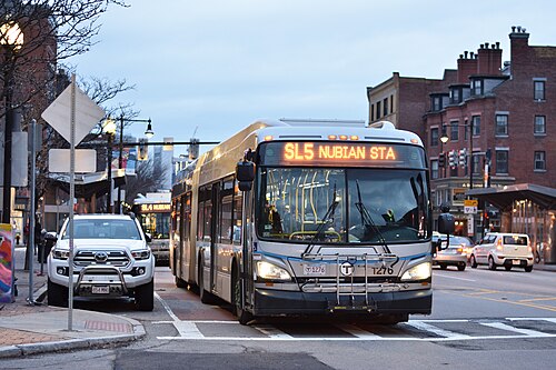 A Silver Line bus signed for Nubian Station stopped on Washington Street, brick rowhouses with paired chimneys behind it in the evening light.