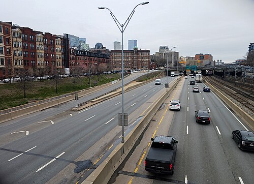 Looking down from the Berkeley Street overpass at the Massachusetts Turnpike, brick rowhouses and a taller mill building lining the north side of the highway cut, the downtown skyline beyond.