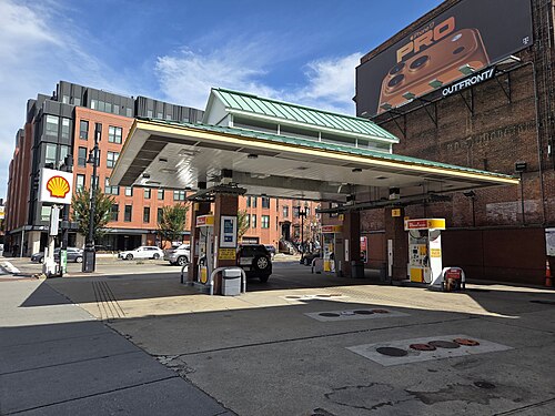 A Shell station under a peaked canopy at Columbus and Massachusetts Avenues, a brick apartment building and a bowfront rowhouse block behind it, a billboard on the brick wall to the right.