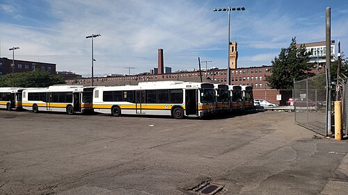 MBTA buses parked in the Albany Street yard, a brick mill building and its chimney stack visible behind the chain link fence.