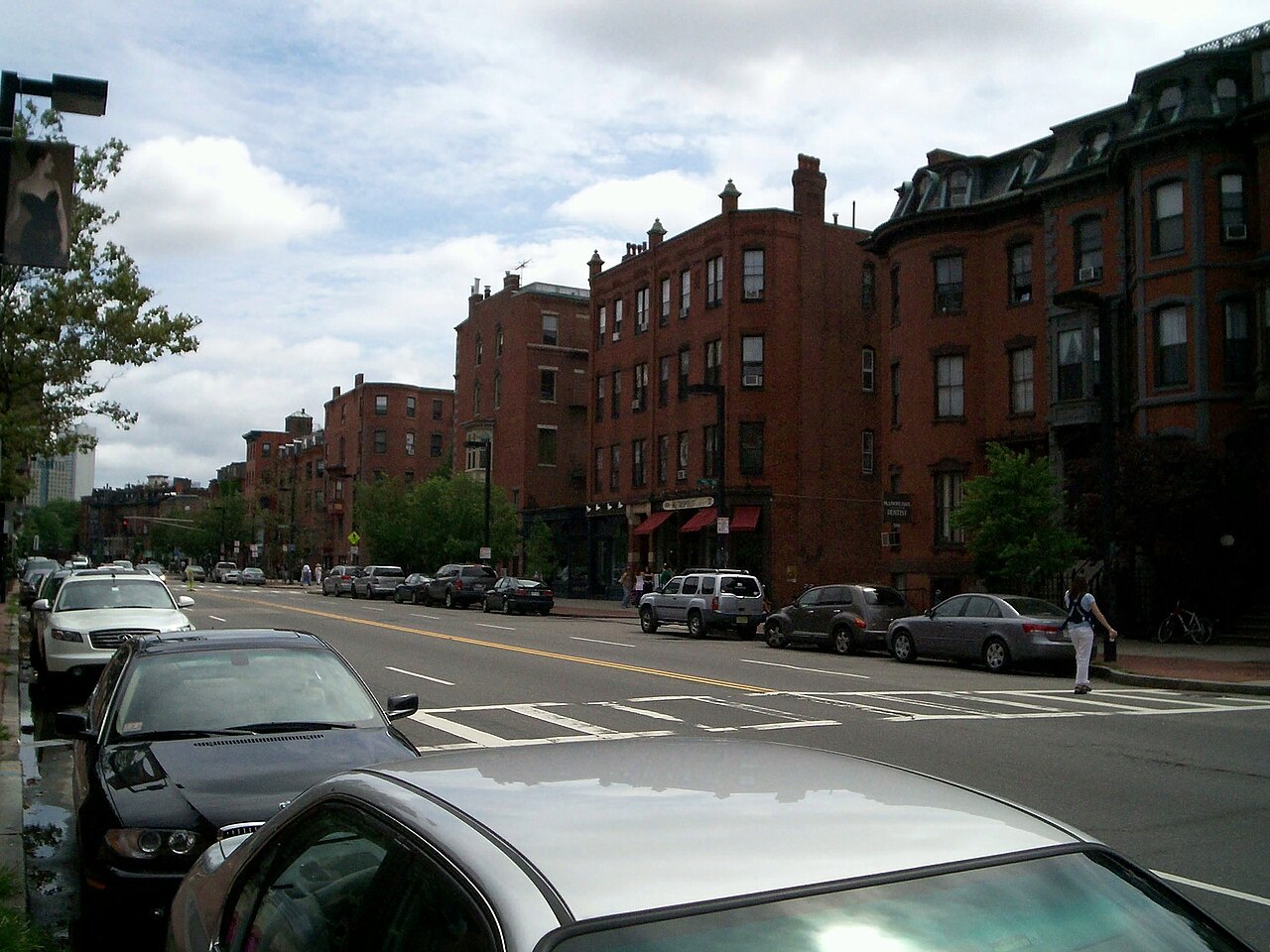 A wide view down Clarendon Street toward Tremont Street, four and five storey brick and brownstone bowfronts lining both sides, storefronts and cafe awnings at the corner, cars parked along the curb.