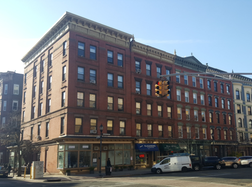 A five storey red brick corner block on Washington Street, ground floor storefronts including a cleaners under striped awnings, a traffic light overhead.