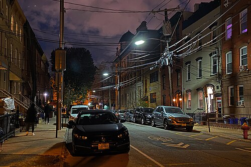 A narrow one way residential street at night, three and four storey brick rowhouses on both sides, cars parked along the curb under wires and a streetlight.