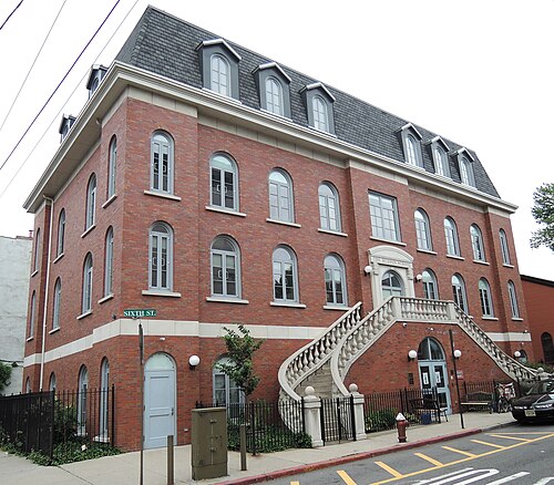 A four storey red brick building at Sixth Street with matching curved stone staircases rising to twin entrances, a mansard roof with dormer windows above.