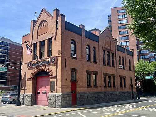 A two storey red brick firehouse on a corner, arched red apparatus doors under a Hoboken Fire Department sign, a stone base along the sidewalk.