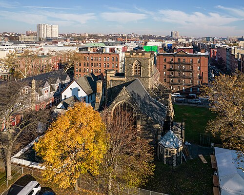 An aerial view of a stone parish church with a peaked slate roof and a lancet window, red brick apartment buildings and fire escapes crowding around it, autumn trees in the foreground.