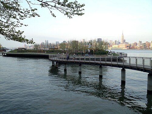 A curved concrete pier extending into the Hudson River toward a small park, the Manhattan skyline including the Empire State Building visible across the water.