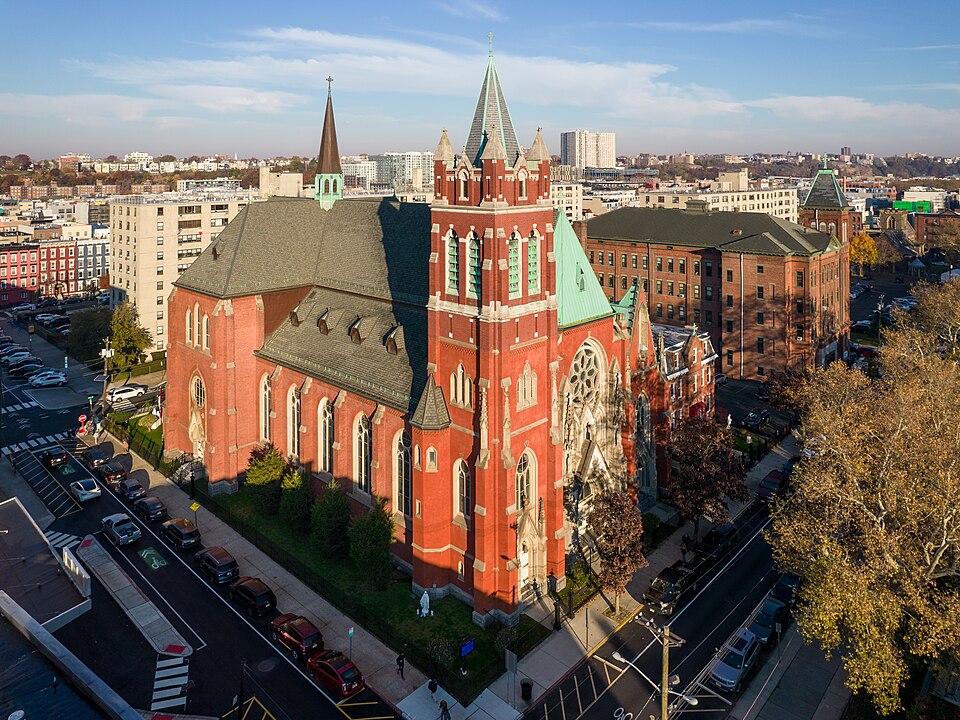 A red brick Gothic Revival church with twin spires and a rose window, facing Church Square, its rectory and parish buildings alongside, seen from above.
