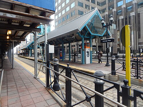 The Hudson-Bergen Light Rail platform at Exchange Place, a peaked blue canopy over ticket machines, office towers and a 101 Hudson Street sign behind.