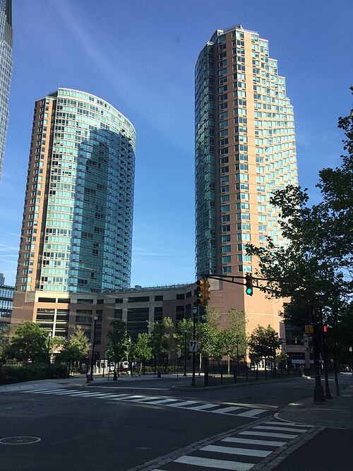 Two curved glass and tan brick residential towers rising over a parking podium at a street intersection near Exchange Place.