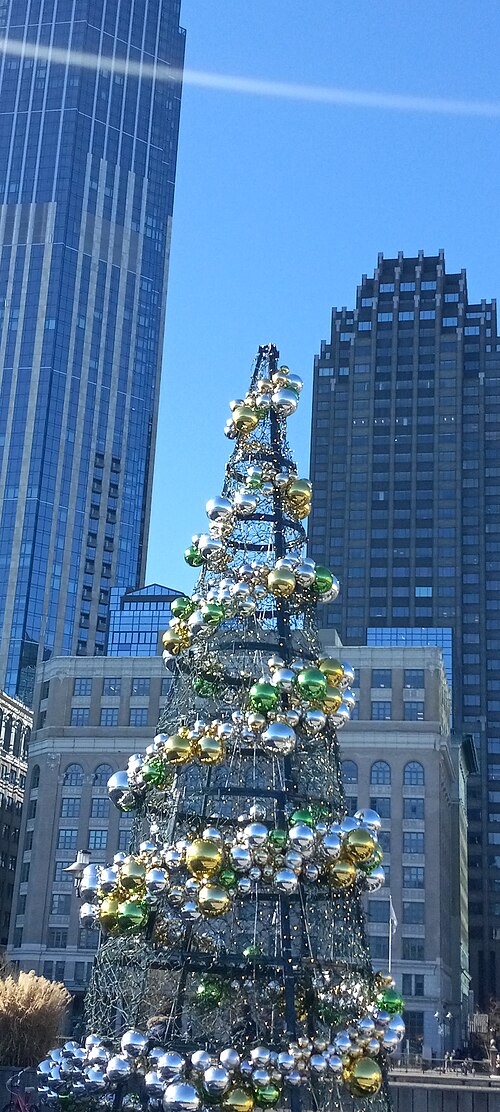 A decorated tree on the plaza, a dark glass tower on one side and a pale stepped crown on the other.