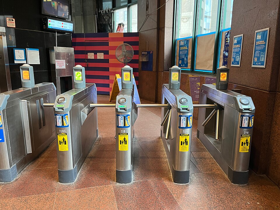 Stainless steel turnstiles inside the Exchange Place PATH station, red stone tile floor, a fan and station signage behind.