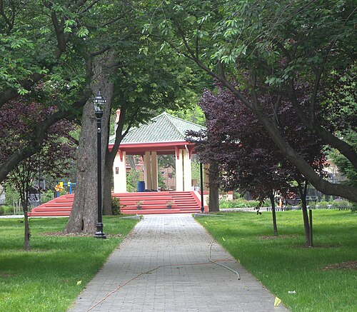 A paved path through the park lawn toward the bandstand, its red steps and green tile roof visible through the trees, seen past a chain link fence.