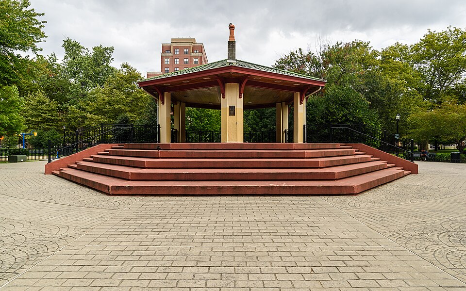 A symmetrical front view of the park's octagonal bandstand, red tiered steps and a green tile roof, lawn and trees around it.