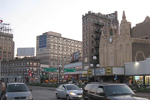 Bergen Avenue and Kennedy Boulevard at dusk, a tall brick office building beside the Loew's Jersey Theatre's clock tower and marquee, traffic crossing the intersection.