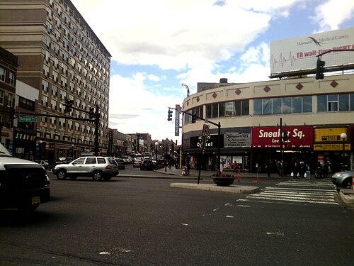 The curved corner of a mid century commercial building at Bergen Avenue, Sneaker Sq and an optometrist's storefront under striped awnings, cars stopped at the crosswalk.