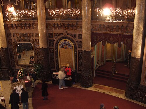 The theatre's lobby from the mezzanine, marble columns, a lit chandelier and a curving staircase down to the carpeted floor.
