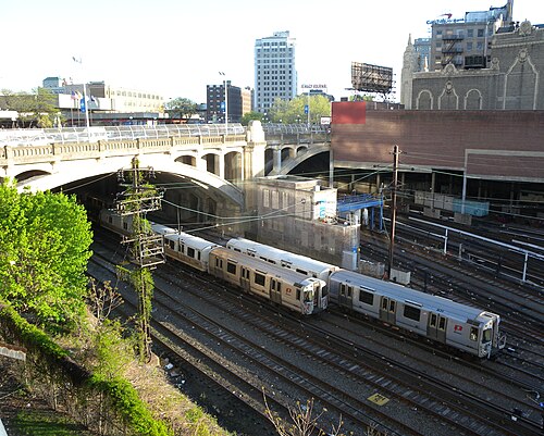 PATH trains sitting on open tracks beneath an arched concrete road bridge, the Jersey Journal sign and the theatre's spire visible on the skyline behind.