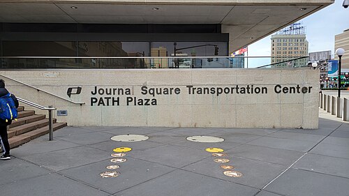The Journal Square Transportation Center PATH Plaza sign carved into a stone wall at the station entrance, with tactile paving leading to the stairs.