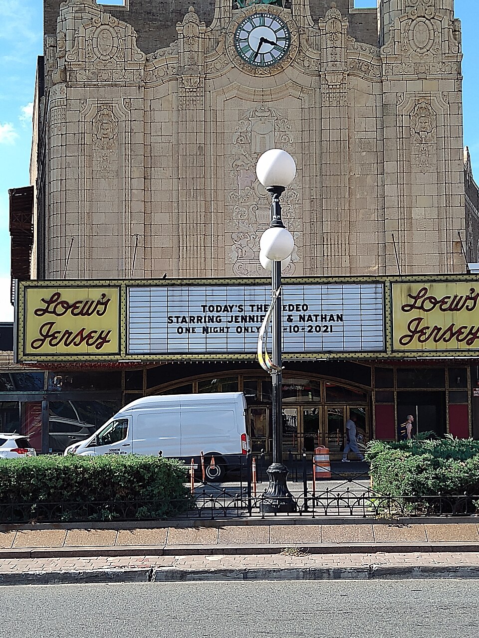 The theatre's marquee reading Loew's Jersey under the clock tower, a double globe lamppost and a parked van at the curb.