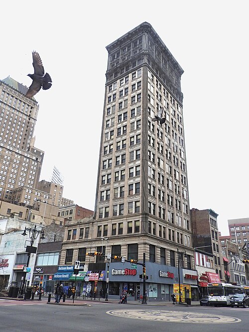 A narrow, columned early skyscraper at Broad and Market, ground floor storefronts and a bus stopped at the corner.