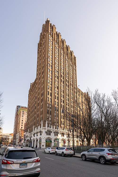 An Art Deco brick tower with a stepped, pointed crown, mature trees fronting it along Broad Street.