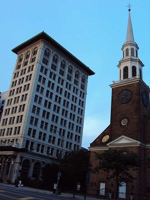 A tall stone office building beside a brick church tower with a clock and a white steeple.