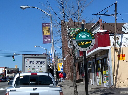 A round green and gold Forest Hill welcome sign on a pole over the sidewalk on Bloomfield Avenue, storefronts and a delivery truck behind it.
